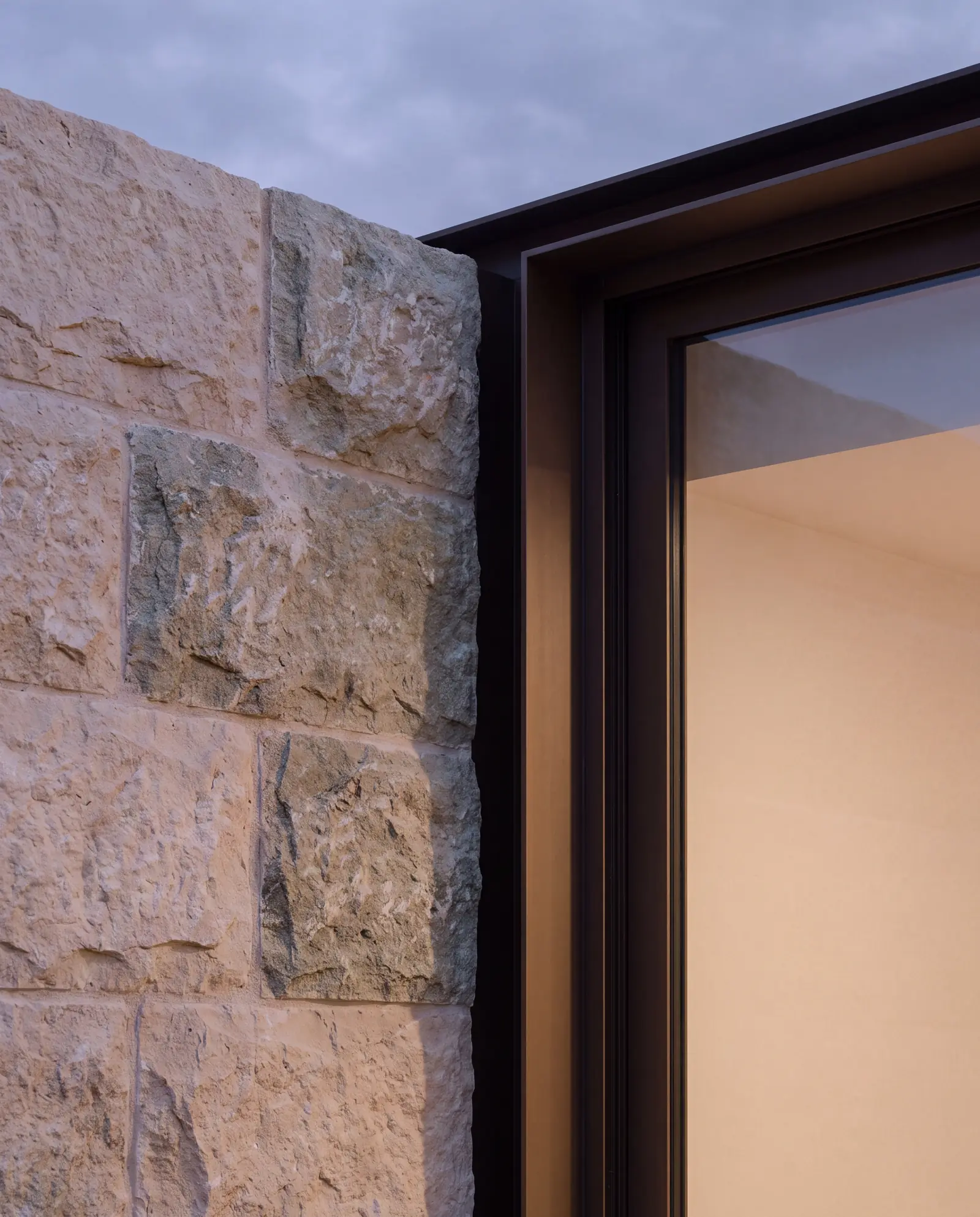 Close view of a coursed limestone wall meeting a slim bronze window frame at dusk